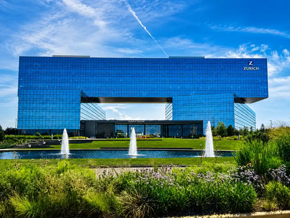 Modern Zurich North America headquarters featuring a striking blue-glass office building, landscaped gardens, reflecting pool with fountains, and a clear blue sky in Schaumburg, Illinois.